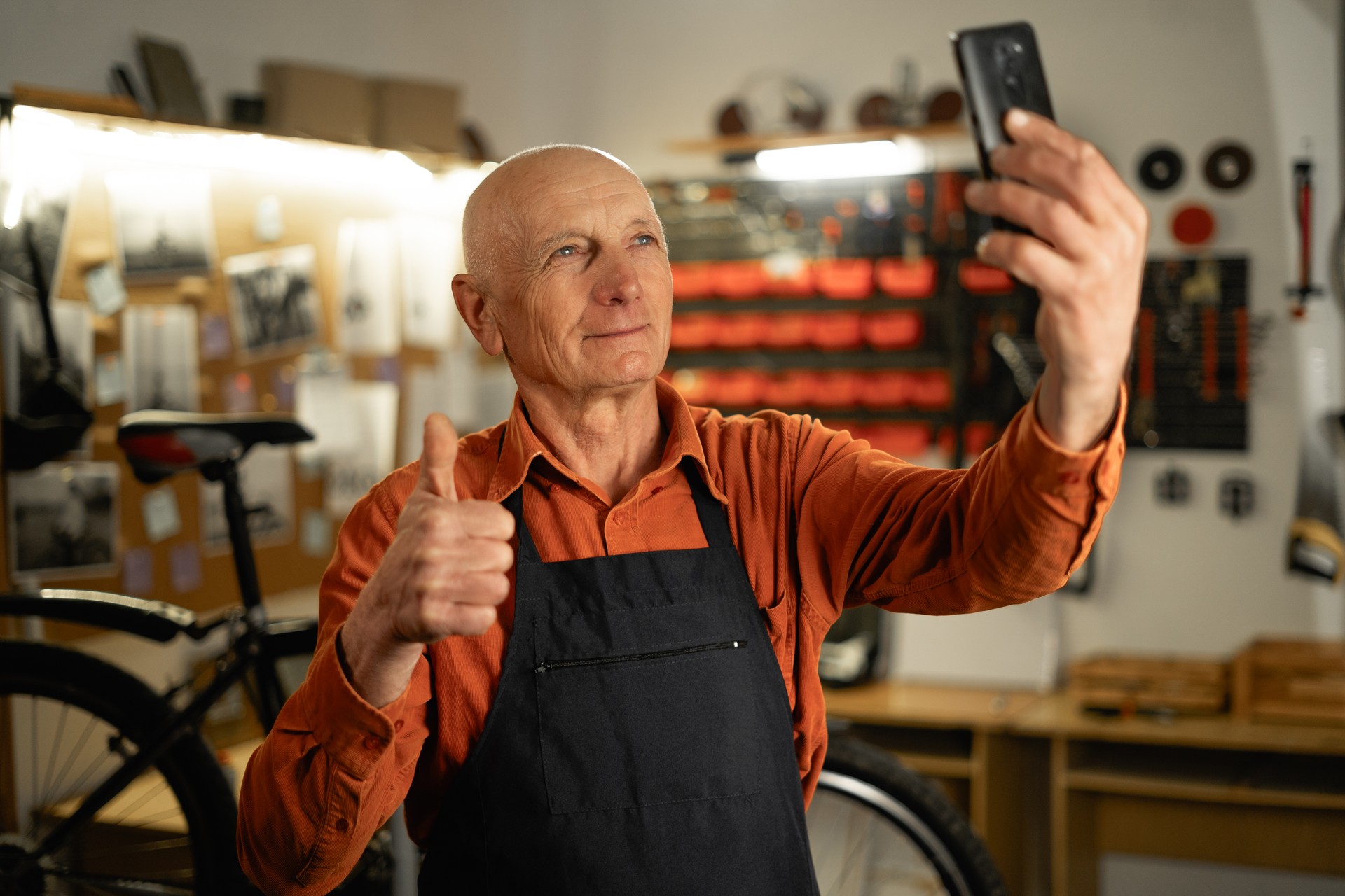 Old male mechanic pausing to take selfie on his mobile phone, repairing or servicing bike in a workshop